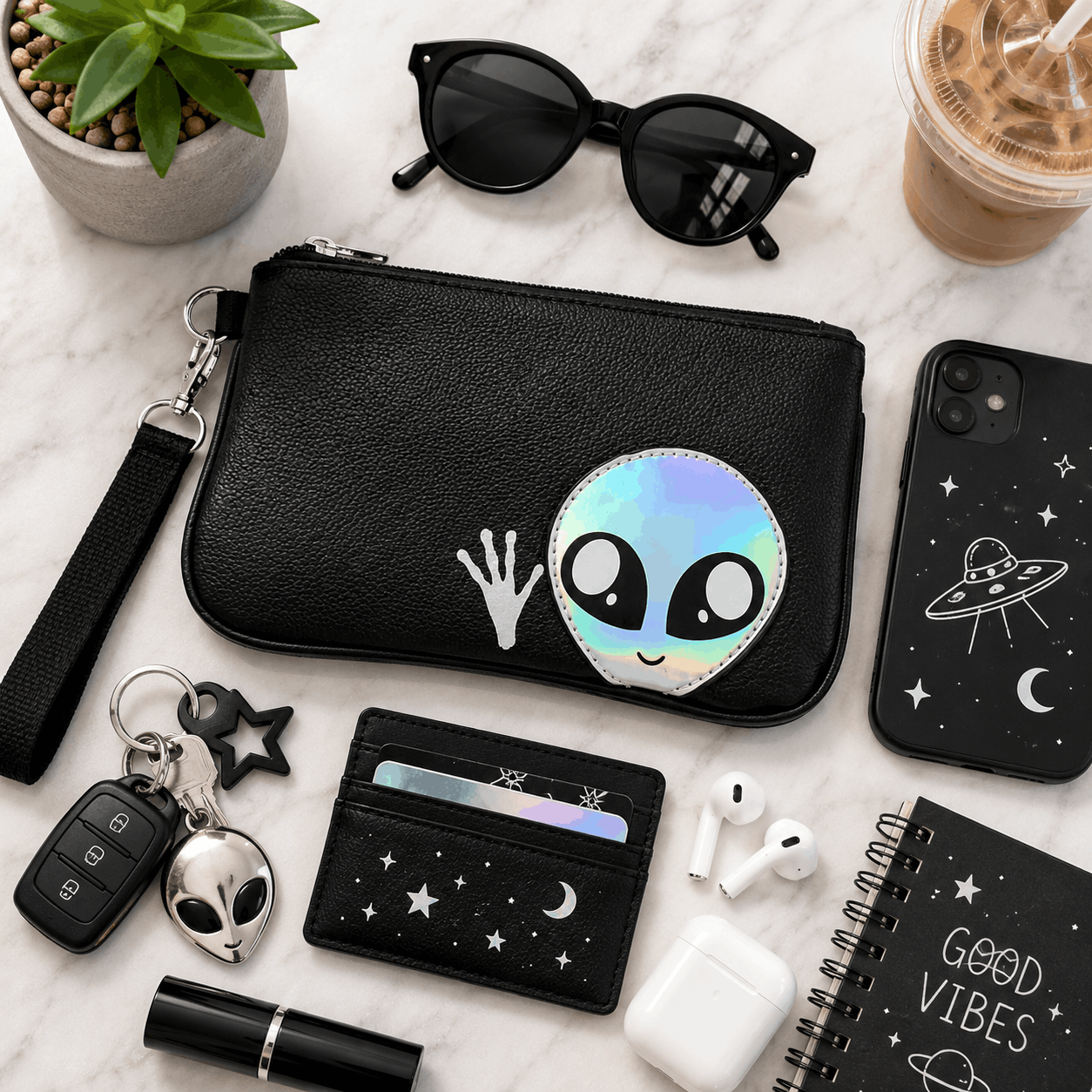 Flat lay of a black alien wristlet with sunglasses, phone, keys, wallet, earbuds, and everyday essentials.