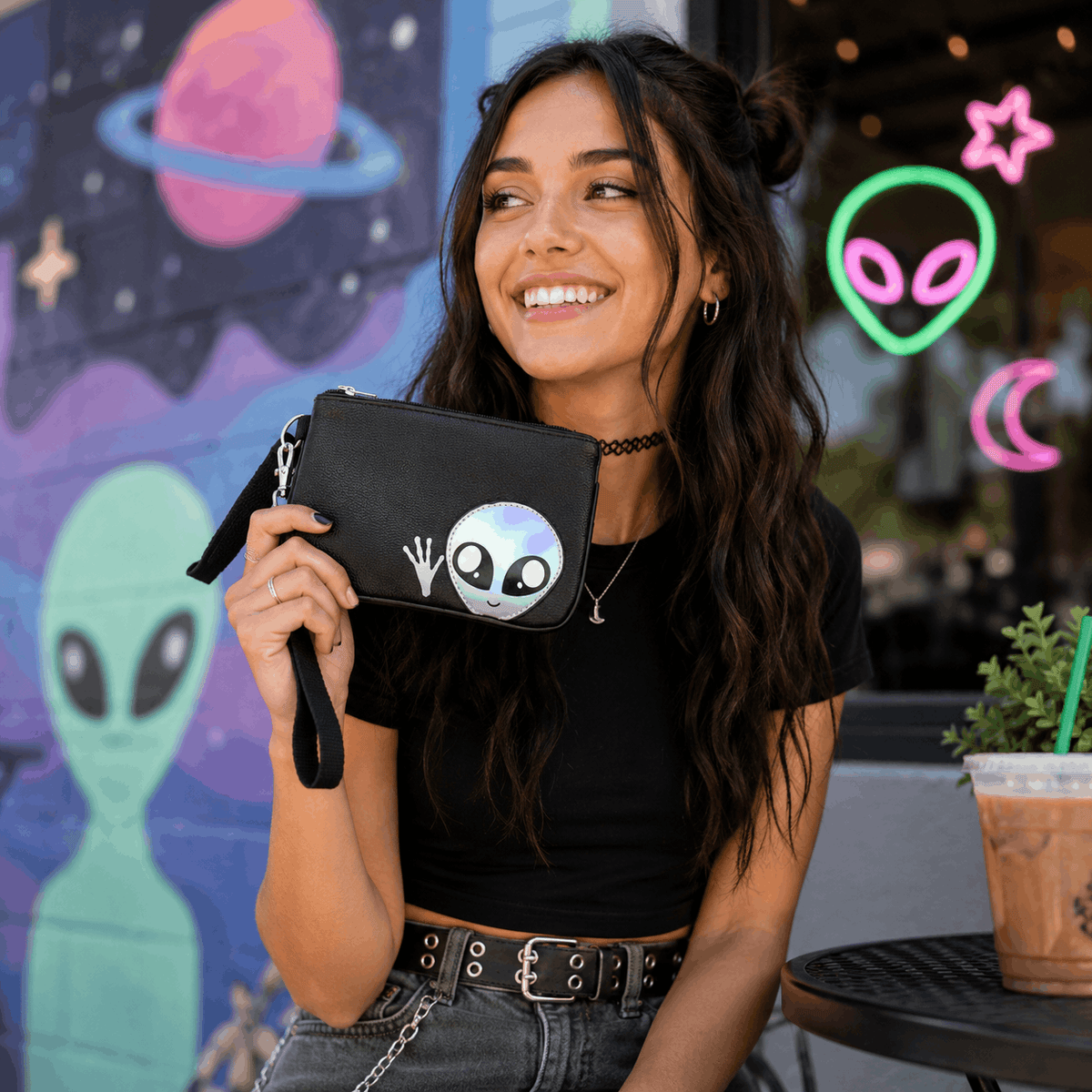 Woman holding a cute black alien wristlet with holographic alien design while enjoying coffee outside.