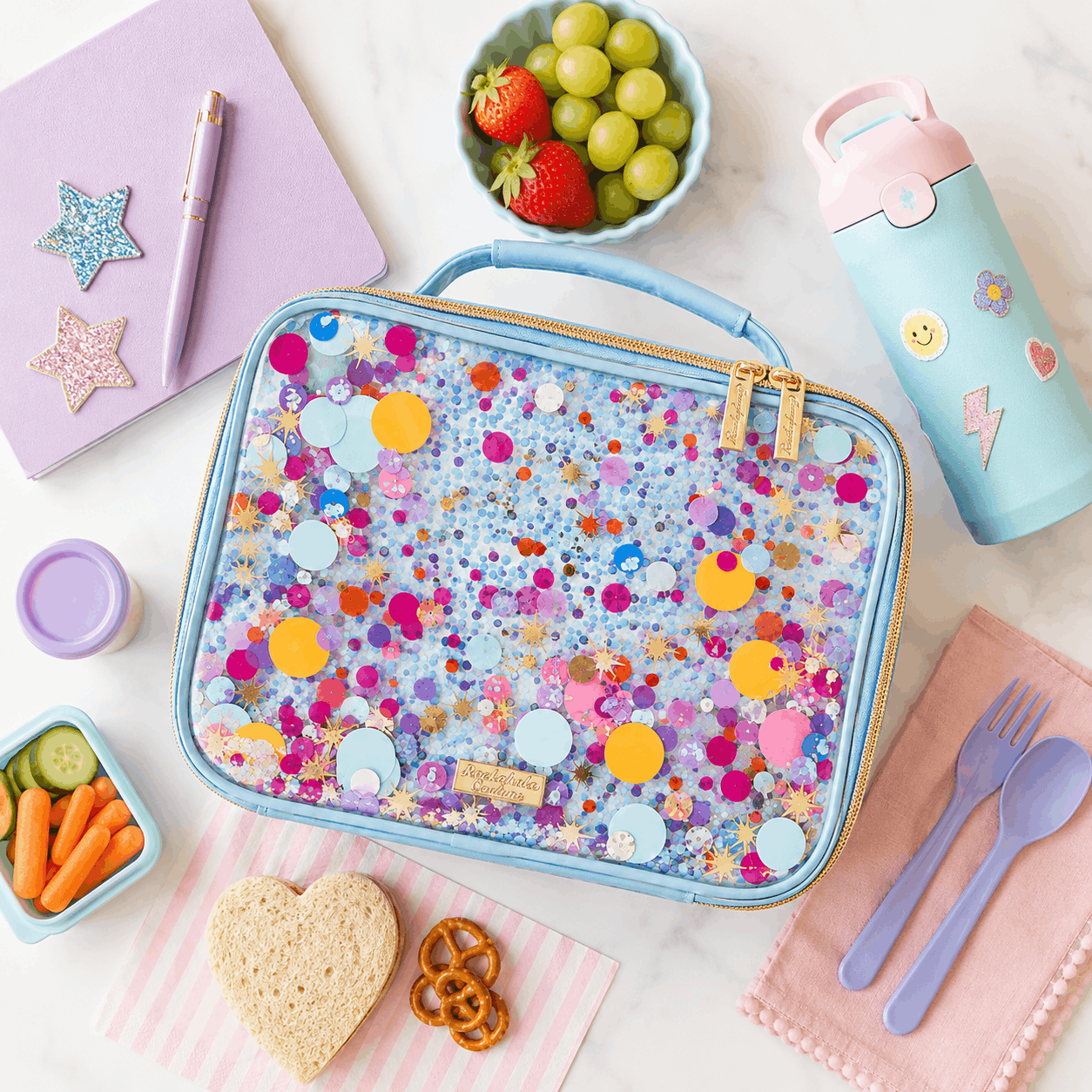 Multicolor confetti insulated lunch box arranged in a flat lay with a water bottle, notebook, fruit, vegetables, sandwich, and school lunch accessories on a light background.